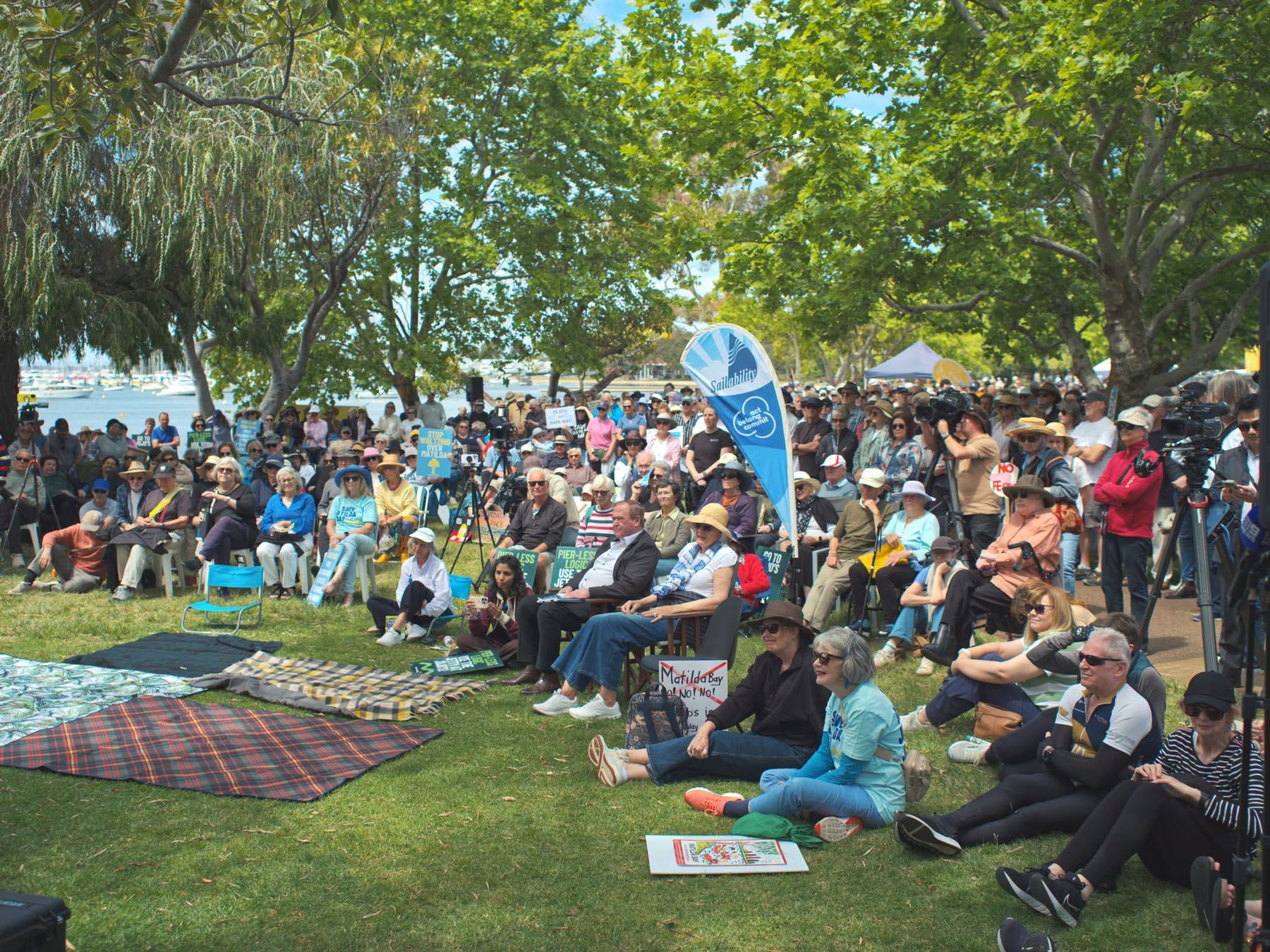 Hundreds of people seated under the trees at the Matilda Bay foreshore during the 18 October Community Action Day, with Sailability flags and Go JoJo's signs.